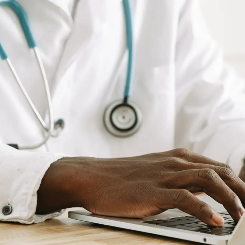 A photo of a male doctor at a laptop, cropped so that only his hand and stethoscope are visible.