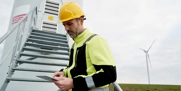 A technician inspects windmill, holding a tablet in his hands.